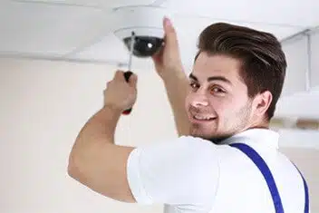 A technician installs a ceiling security camera, smiling confidently while using a screwdriver in a well-lit indoor setting.